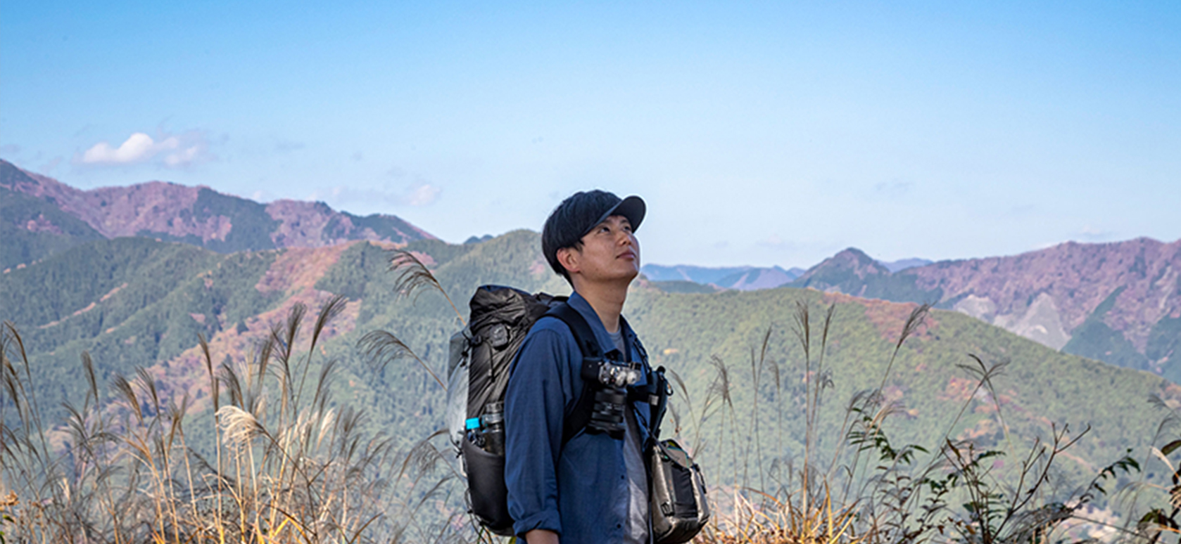 すすきが生えた山頂で青空を見上げるJINさん